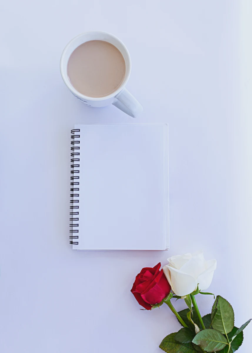 Açık renkli zeminde boş beyaz spiral defter, kahve fincanı, kırmızı ve beyaz güller.